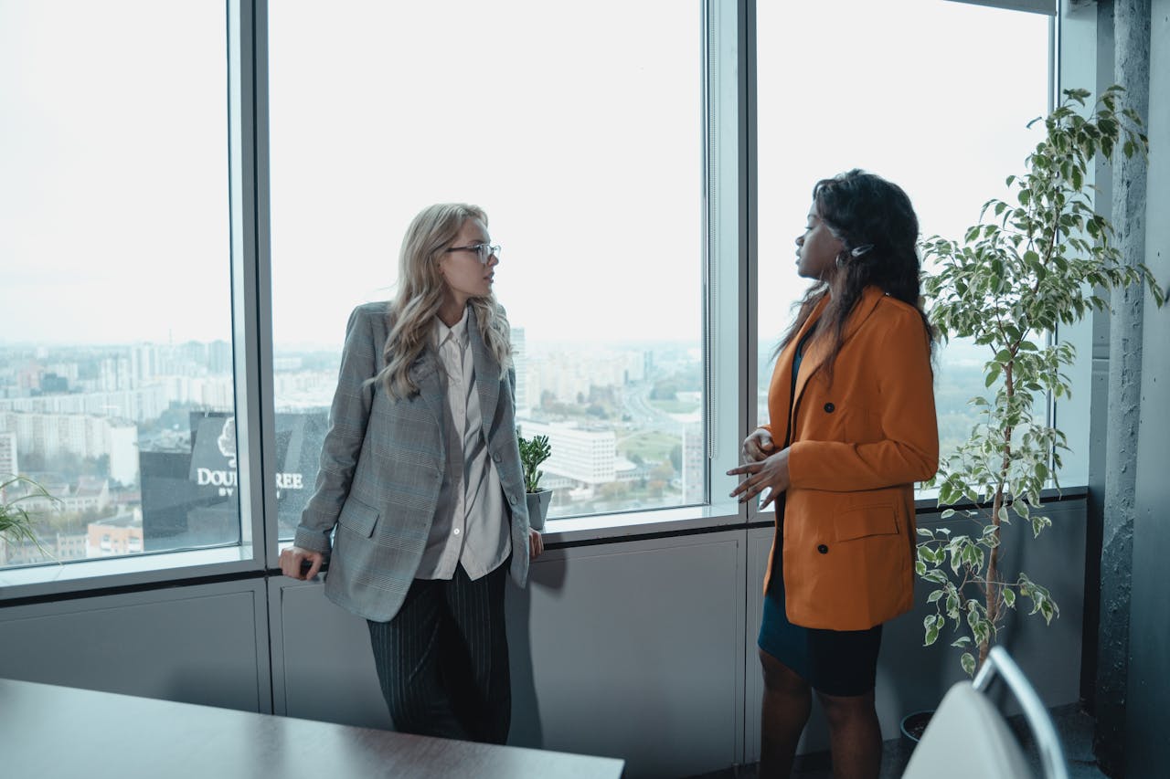 Two businesswomen conversing in a modern office with a city view, promoting corporate diversity.