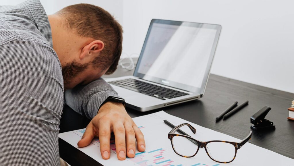 Exhausted man resting at desk with laptop, glasses, and charts indoors.