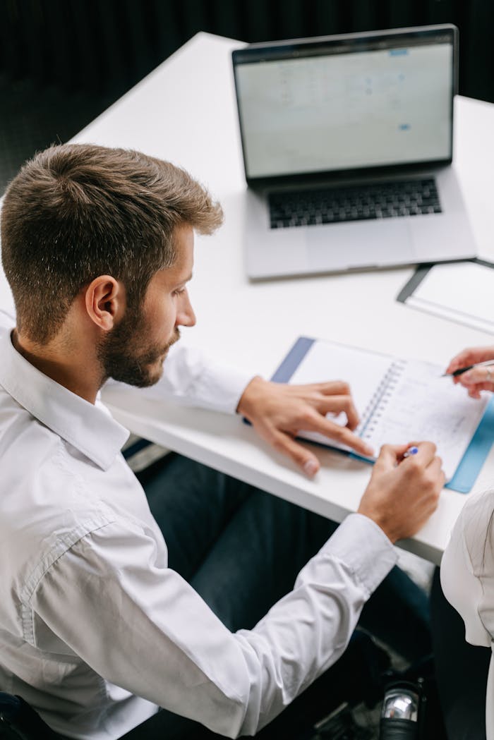 Man writing in a notebook on a desk with a laptop open in an office environment.