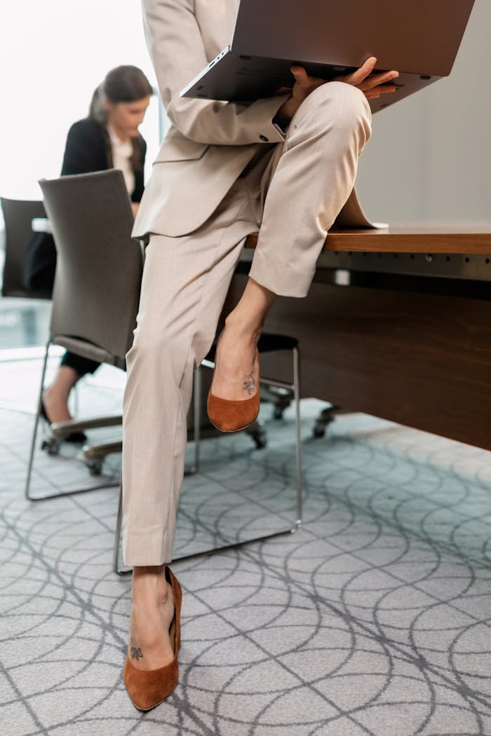 Businesswoman in a tan suit sitting on a desk, working on a laptop in a modern office.