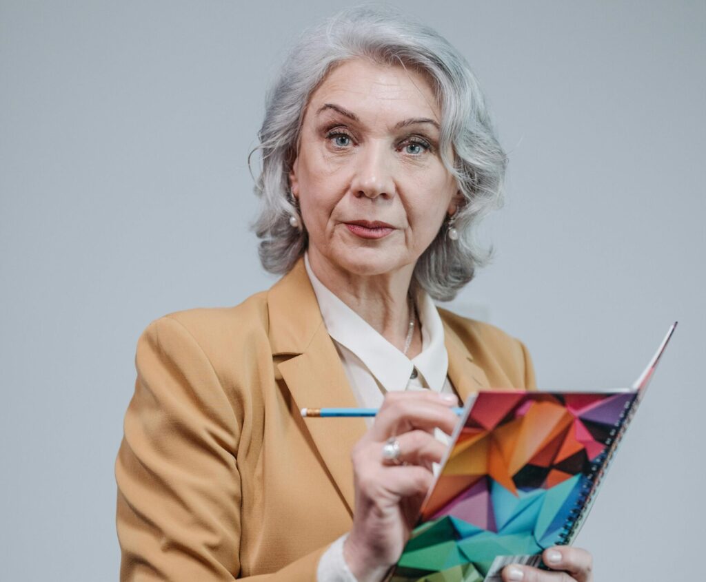 Elderly woman in brown blazer holding a colorful notebook against a gray background.